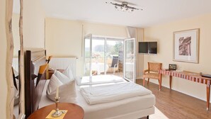 A hotel room with a bed, a table, a chair, a TV, and a balcony with a view. - Seebauer Hotel Gut Wildbad (Wemding)