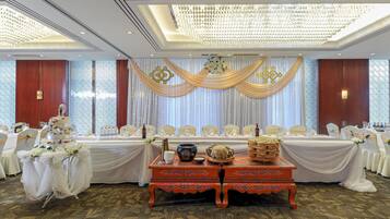 A banquet hall with a long table set for a formal event, draped with white and gold fabric, and a decorative ceiling light fixture.