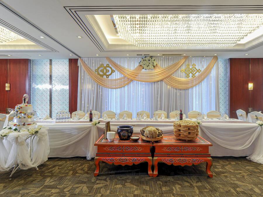 A banquet hall with a long table set for a formal event, draped with white and gold fabric, and a decorative ceiling light fixture.