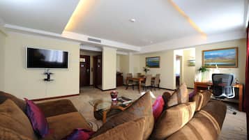 A hotel living room with a flat-screen TV, a sofa, a coffee table, and a dining area with chairs and a table.
