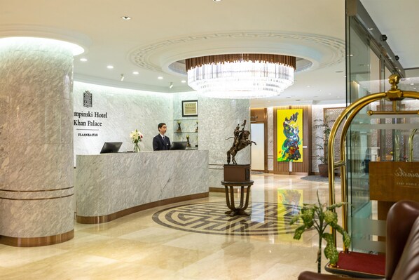A hotel reception area with a marble reception desk, a receptionist, and a chandelier.
