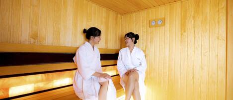 Twee personen in een houten sauna met banken, een klok en een houten plafond.