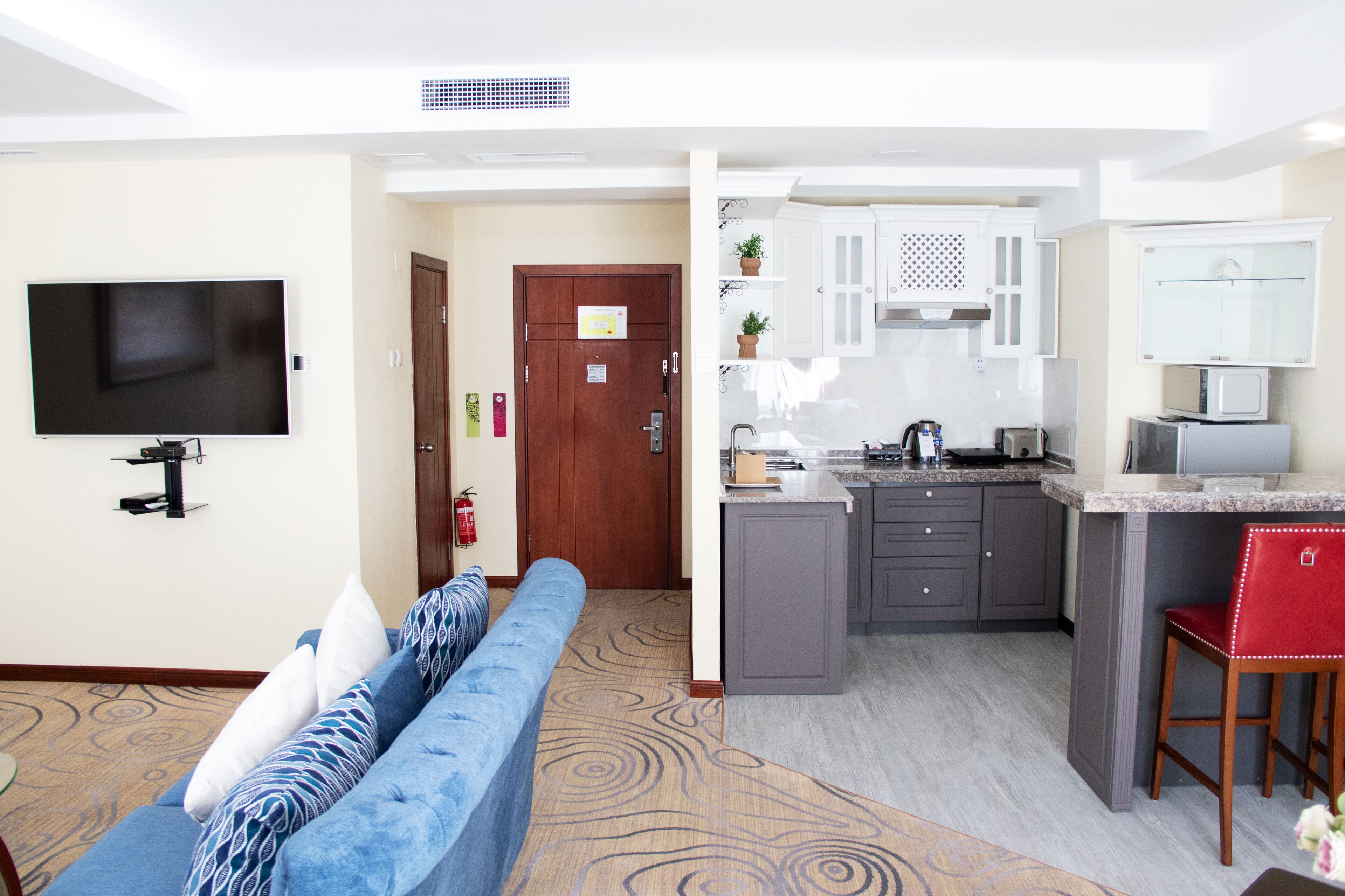 A modern living room with a blue sofa, a flat-screen TV, and a kitchen area with a microwave, toaster, and bar stools.