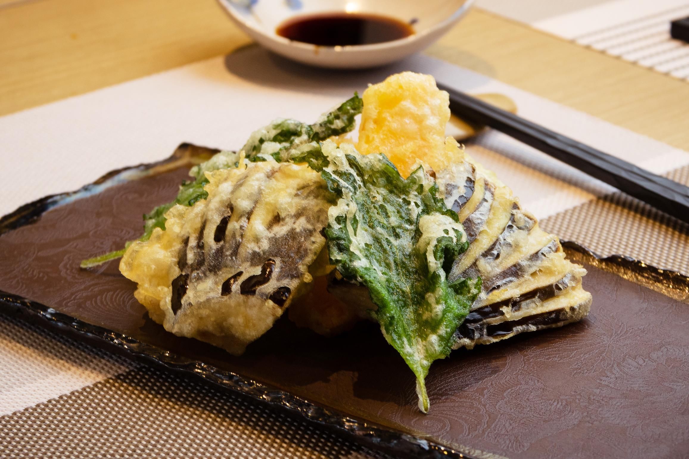 A dish of tempura with a dipping sauce, placed on a dark plate with chopsticks and a bowl of sauce.