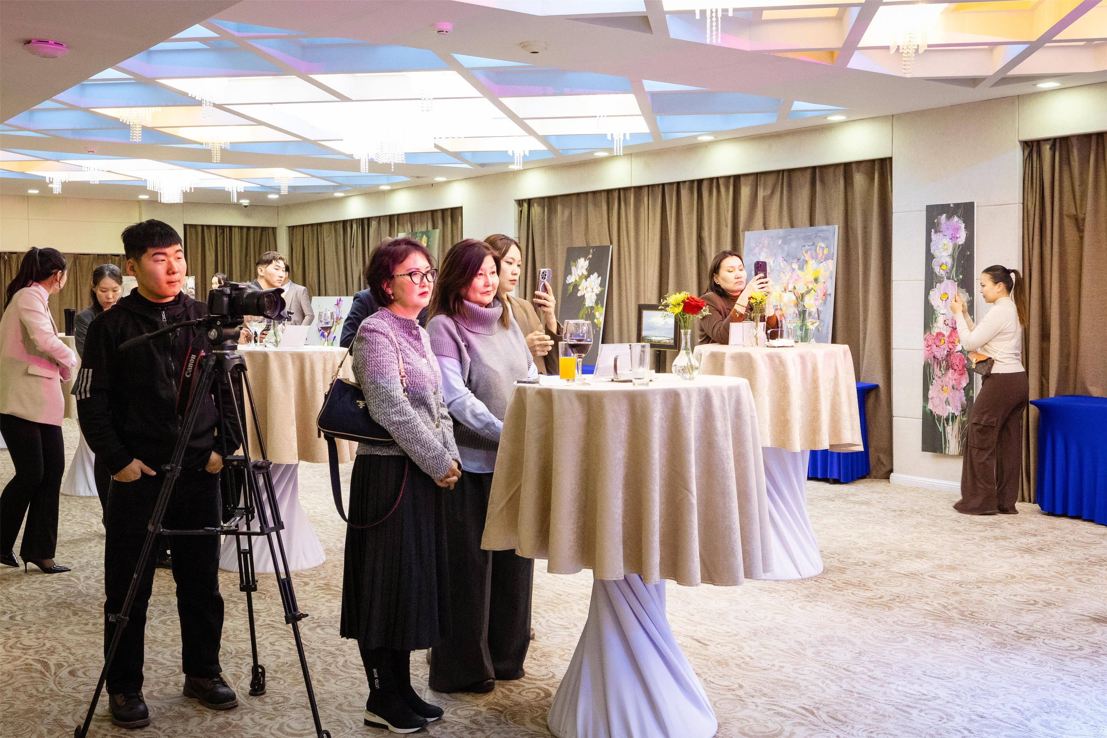 A room with tables set up for a meeting or event, a camera on a tripod, and people engaged in conversation.