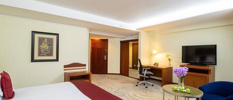 A hotel room with a bed, a desk, a chair, a television, and a small table with a vase.