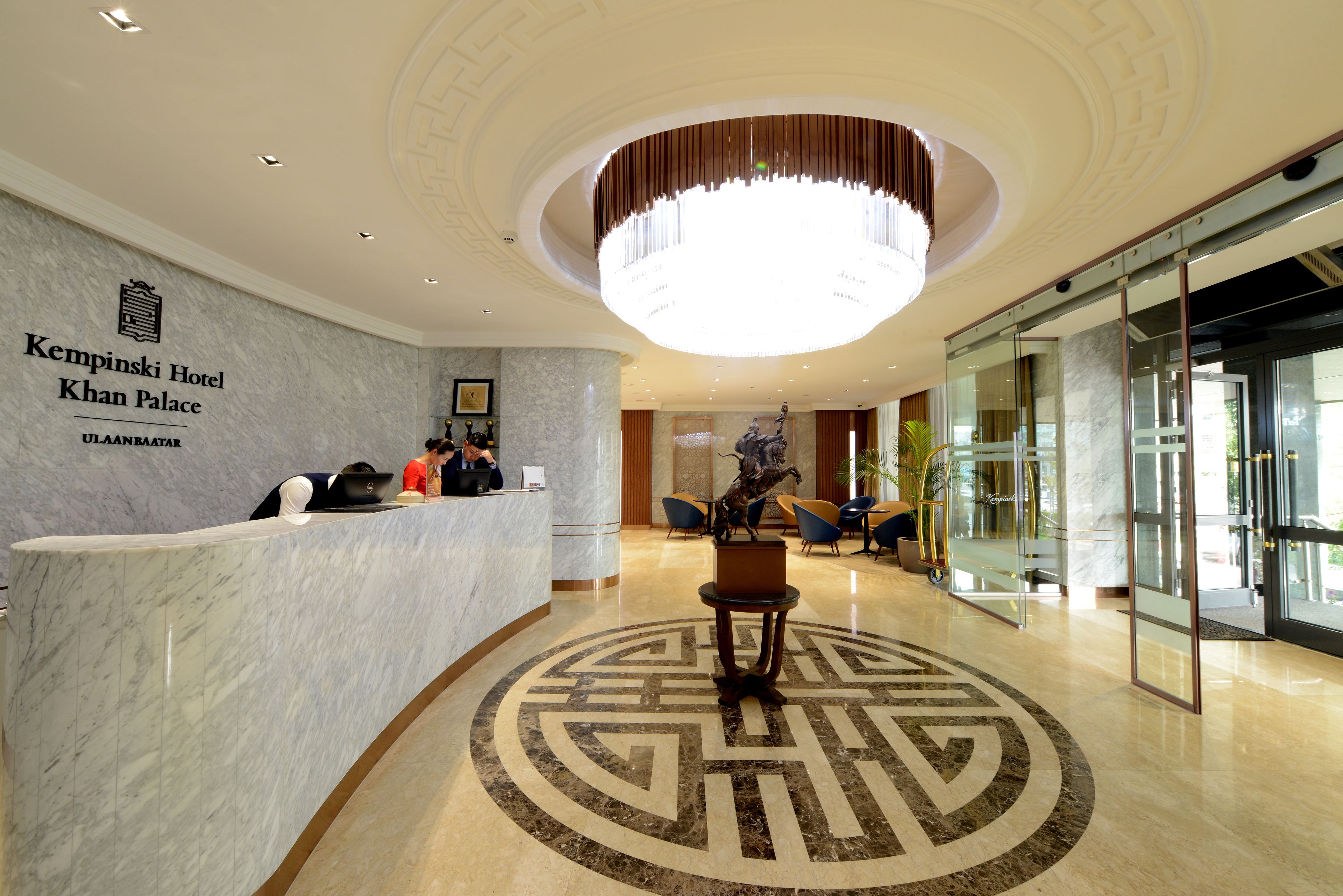 Zona de recepción de un hotel con mostrador de mármol, una lámpara de araña grande y un diseño decorativo en el suelo.