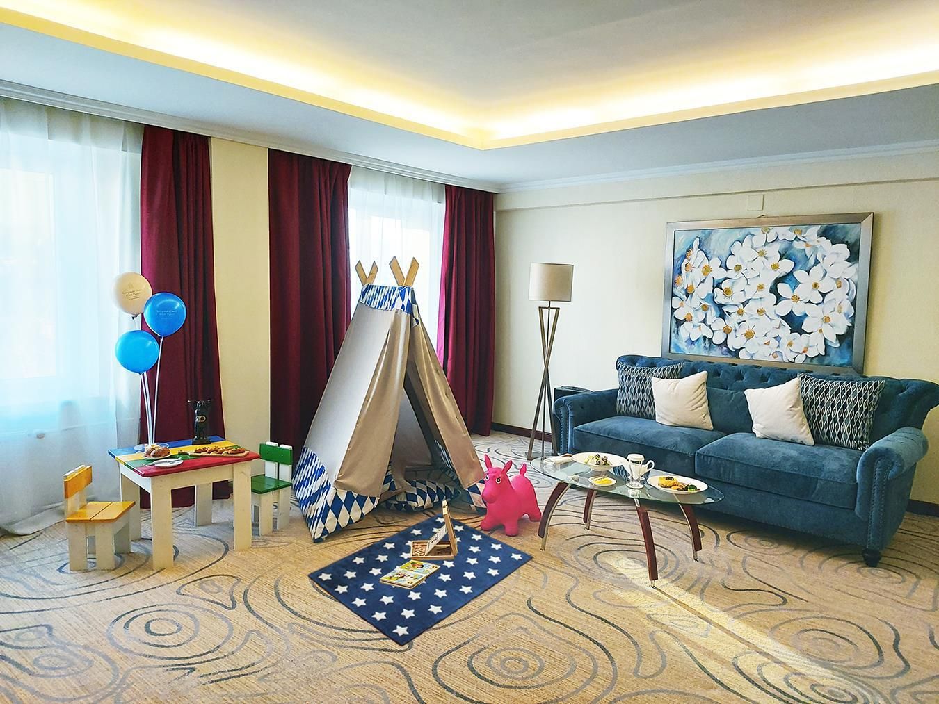 Una habitación de hotel con un sofá azul, un cuadro, un tipi, un tapete con estampado de estrellas y una pequeña mesa con comida y bebidas.