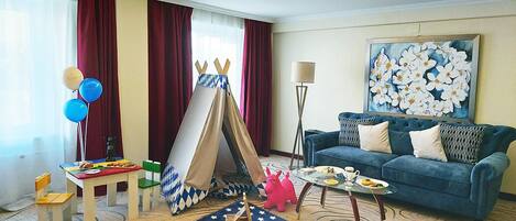 Una habitación de hotel con un sofá azul, un cuadro, un tipi, un tapete con estampado de estrellas y una pequeña mesa con comida y bebidas.
