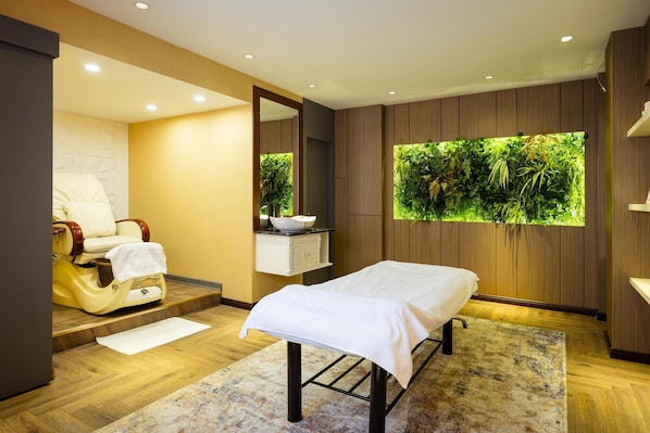 A modern spa room with a massage table, a massage chair, a large window with a view of greenery, and wooden paneling on the walls.