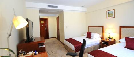 A hotel room with two beds, a desk, a chair, a TV, and a lamp.
