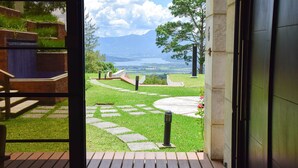 Vista de dentro de um edifício para um jardim ajardinado com um caminho de pedra, um deck de madeira e uma cadeia montanhosa ao longe.