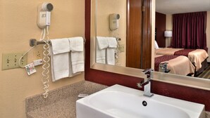 A hotel bathroom with a sink, towel rack, hair dryer, and a mirror reflecting a bedroom with a bed and curtains.
