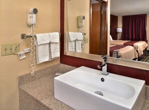 Baño de hotel con lavamanos, perchero para toallas, secador de pelo y un espejo que refleja un dormitorio con cama y cortinas.