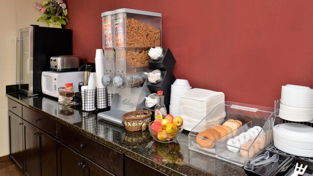 A breakfast bar with a coffee maker, cereal dispenser, and various food items like fruits and pastries.