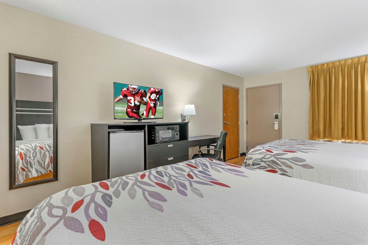 Ein Hotelzimmer mit zwei Betten, einem Schreibtisch, einem Fernseher, einem Kühlschrank und einem Spiegel.