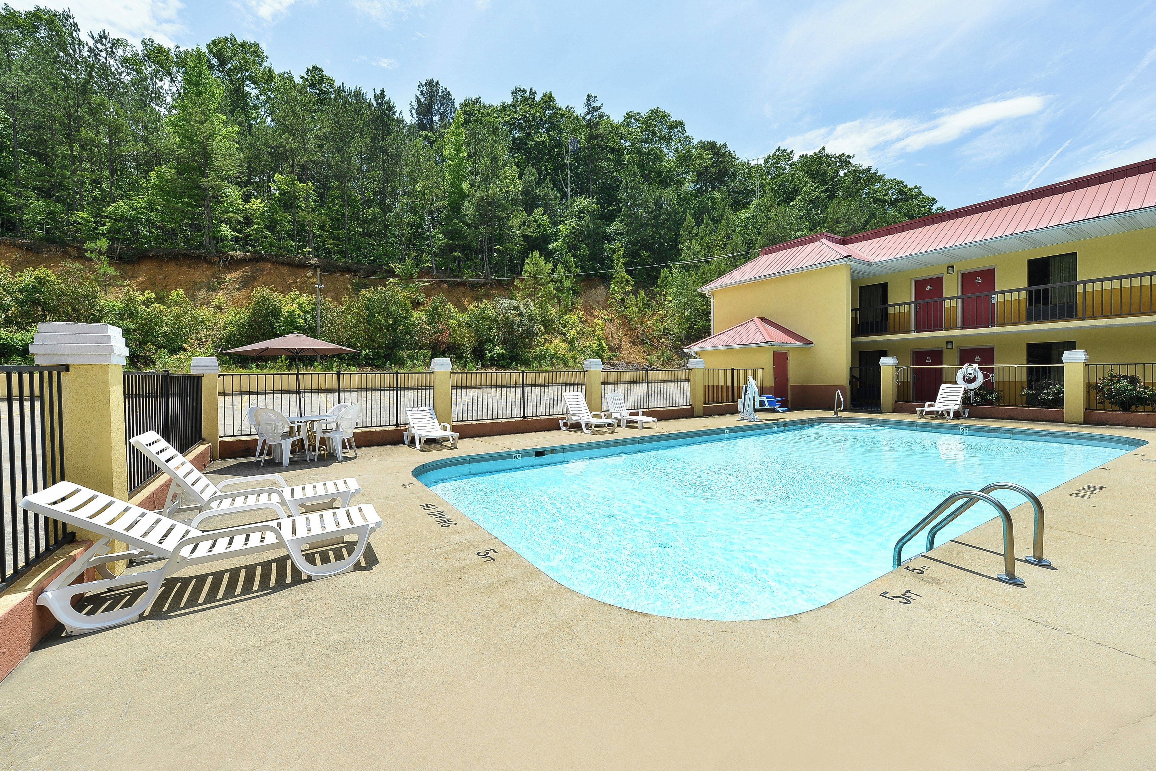 A swimming pool with lounge chairs and umbrellas, surrounded by a fenced area with a building in the background.