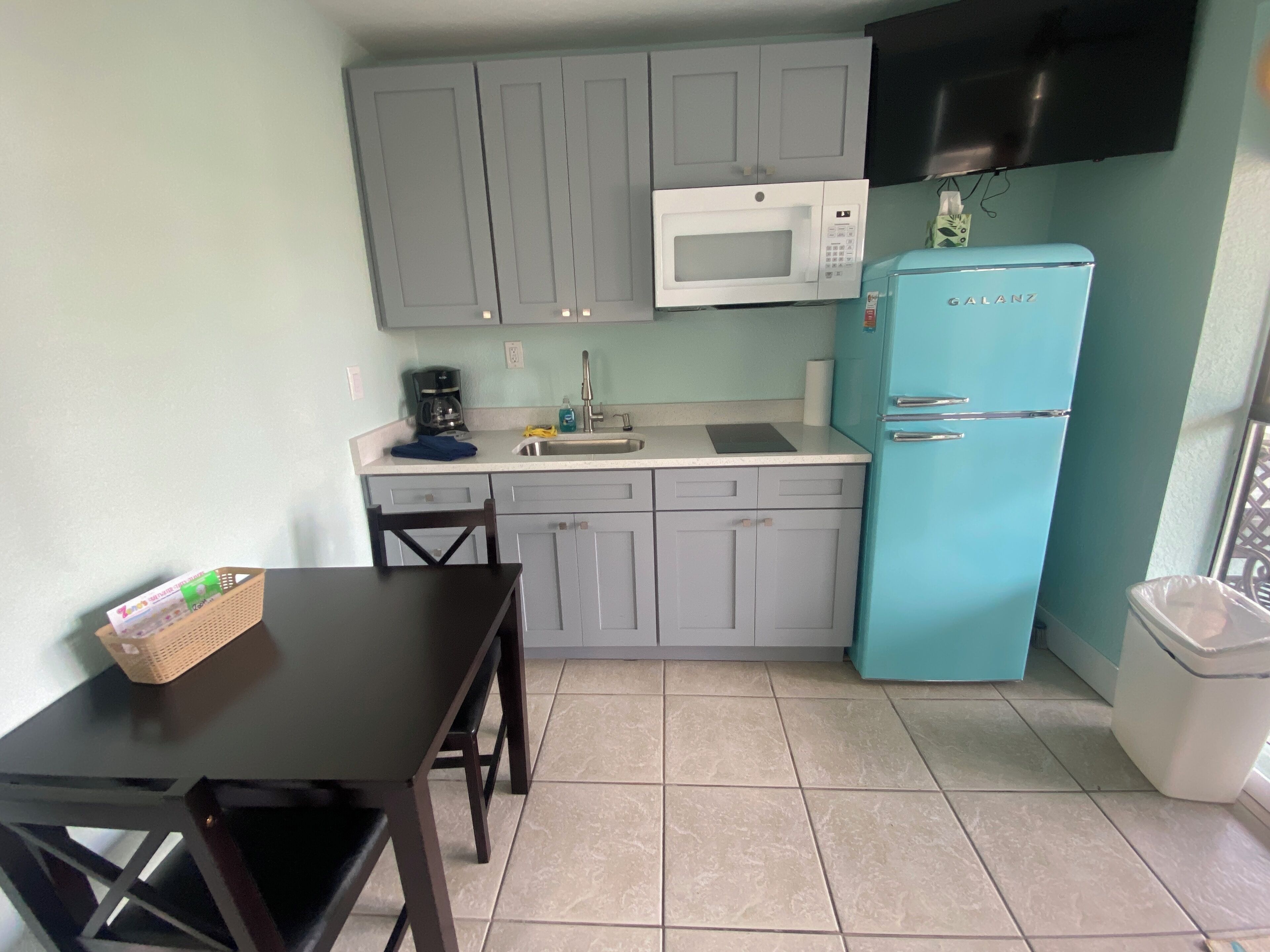 A compact kitchen with grey cabinets, a white microwave, a blue refrigerator, and a small dining table.