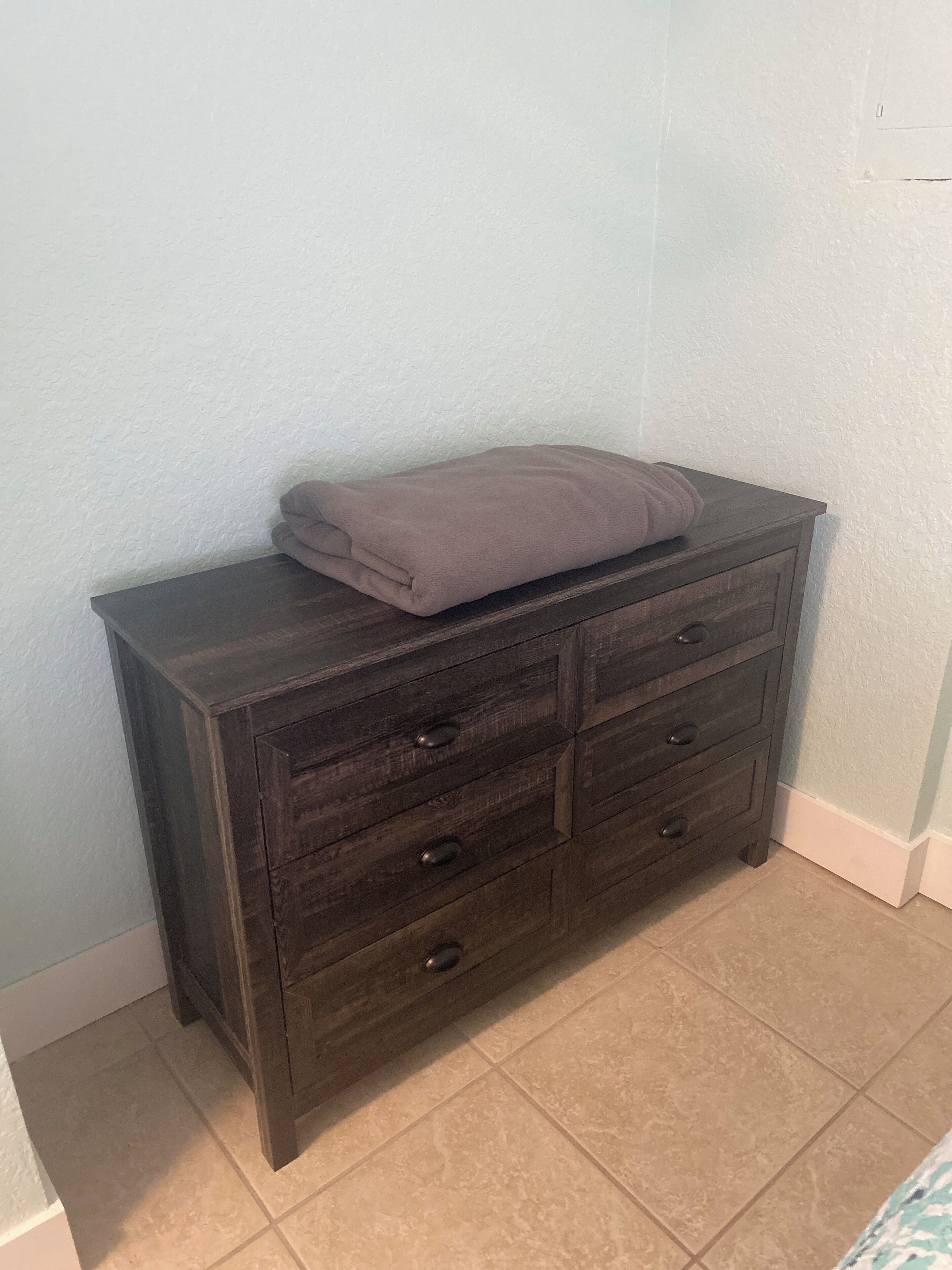 A dark wooden dresser with four drawers and a folded towel on top, positioned against a light blue wall with a tiled floor.