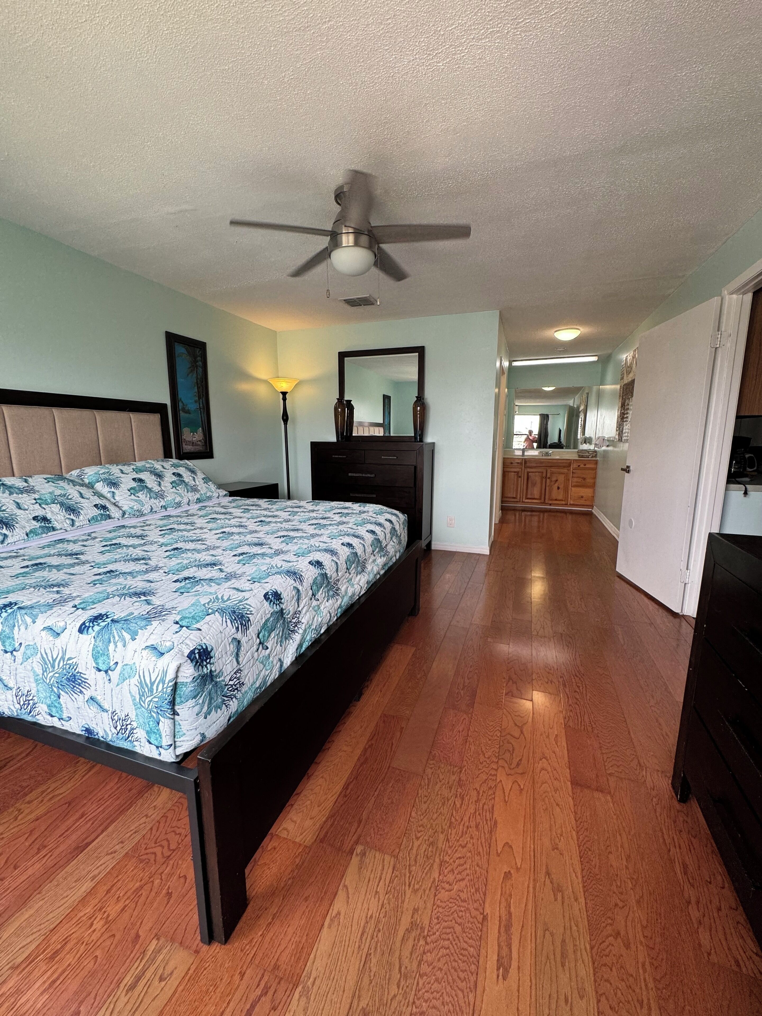 A bedroom with a bed, a dresser, a nightstand, a ceiling fan, and a view into the kitchen.