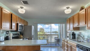 A modern kitchen with wooden cabinets, a central island with bar stools, a large window with a view of the outdoors, and a ceiling fan. - Sand Vista Vacation Rentals (Redington Shores)