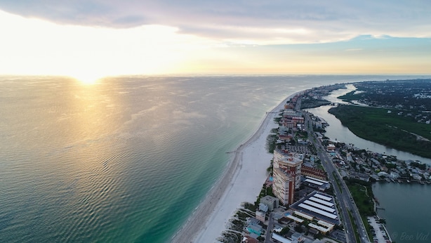 An aerial view of a coastal city with a long beach, a river running parallel to the shore, and a dense urban area with buildings and roads. - Sand Vista Vacation Rentals (Redington Shores)