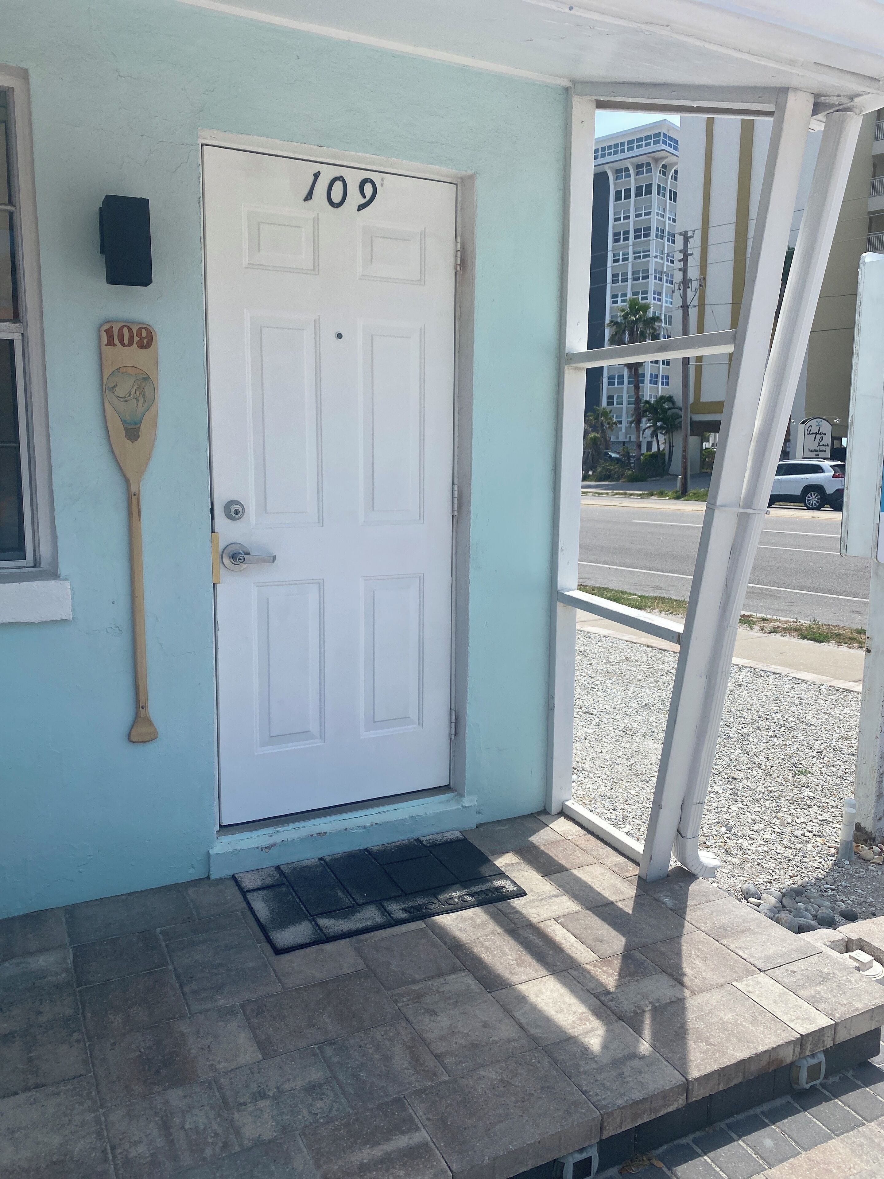 A white door with the number 109, a wooden paddle leaning against the wall, and a view of a street with buildings and cars.