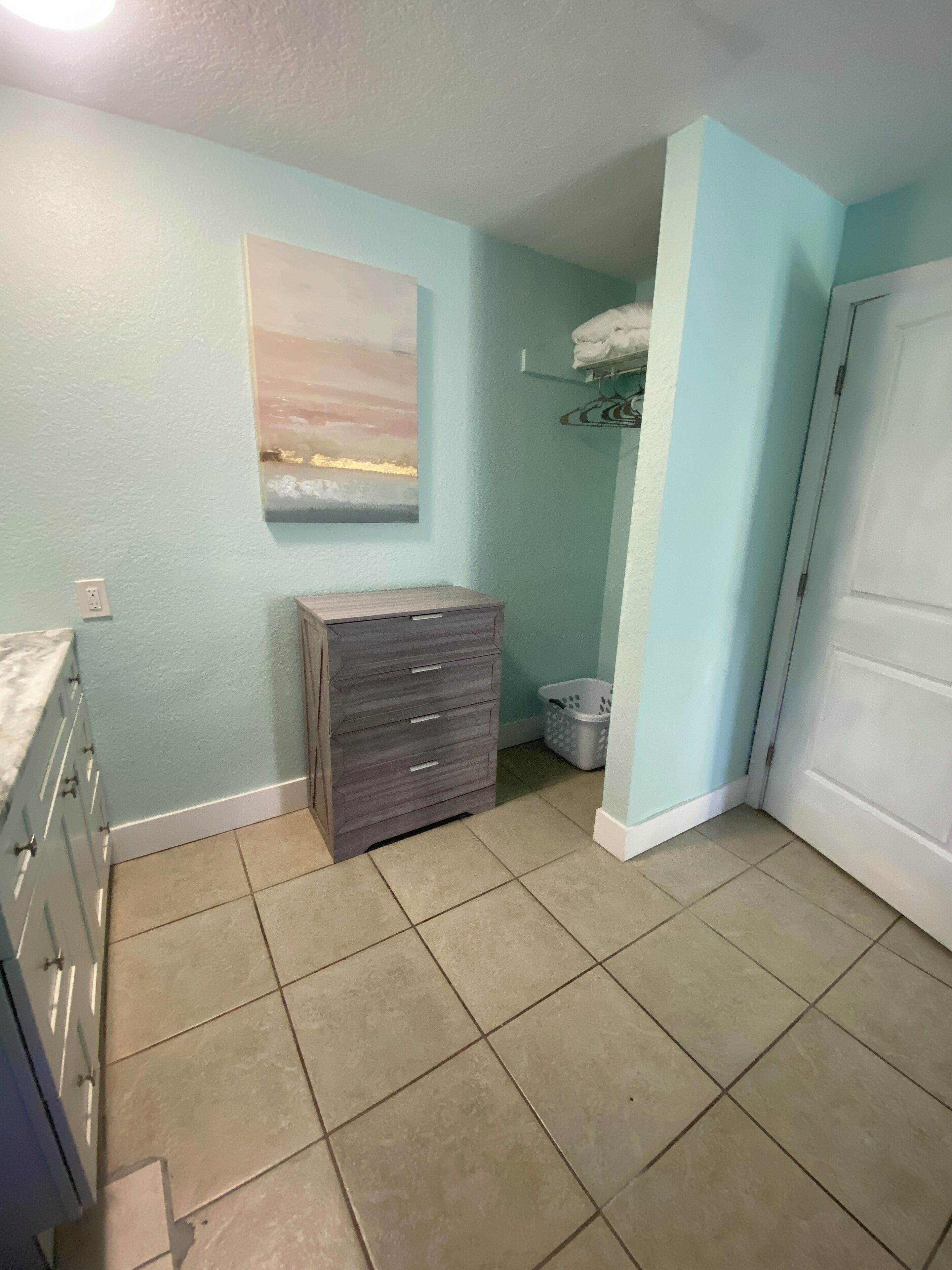 A bathroom with light blue walls, a gray dresser, a white door, and a painting of a landscape.