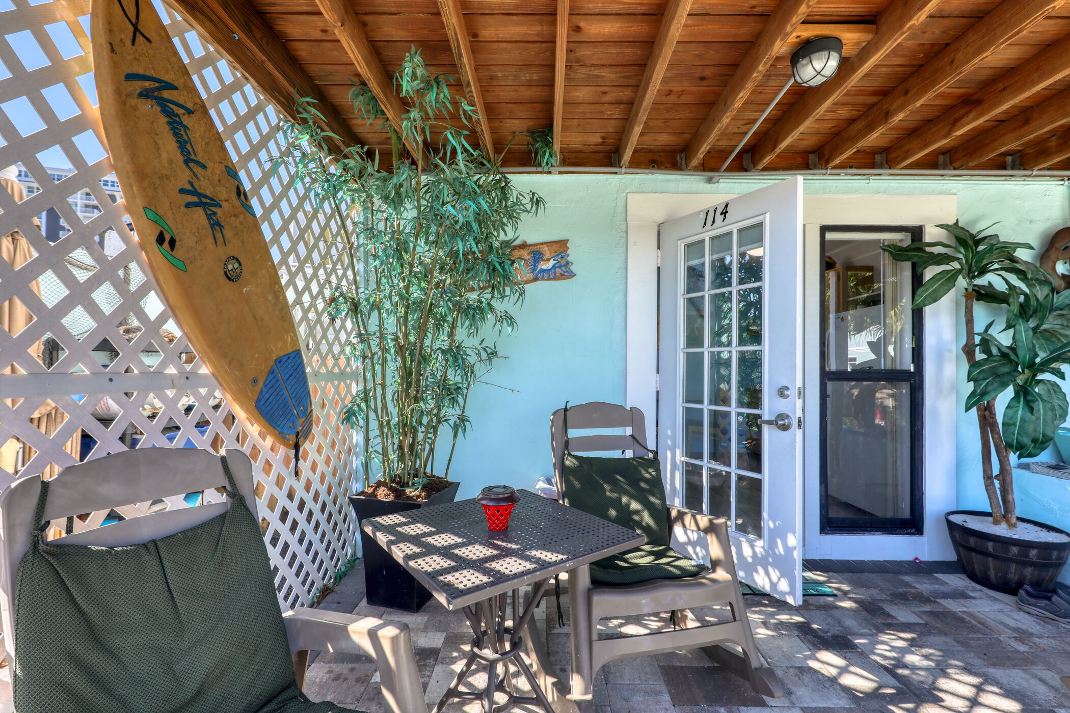 Une terrasse couverte avec une table et des chaises, une planche de surf en bois et des plantes en pot.