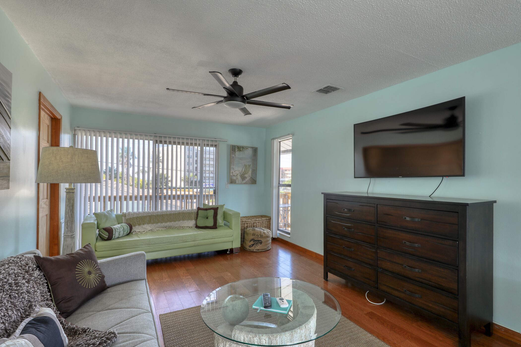 A living room with a green sofa, a glass coffee table, a dark wooden dresser, and a ceiling fan.