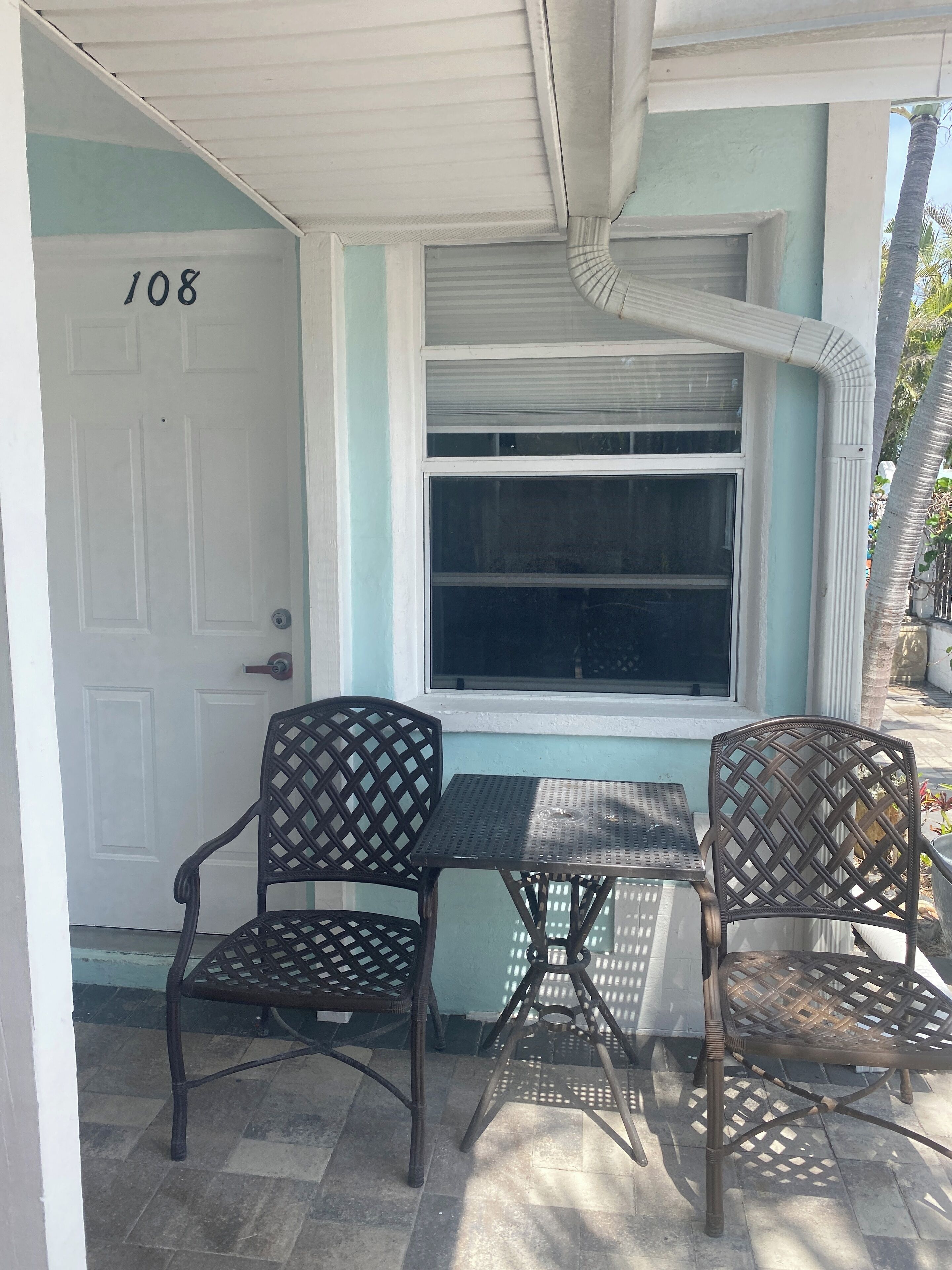 Une véranda avec deux fauteuils en osier et une petite table, une porte blanche portant le numéro 108, et une fenêtre surmontée d’un auvent blanc.