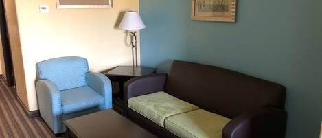 A hotel room with a blue armchair, a brown sofa, a small table, and two framed artworks on the walls.