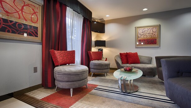 A modern living room with a grey sofa, red cushions, a round ottoman, and a glass coffee table. - Best Western Plus Goodman Inn & Suites (Horn Lake)