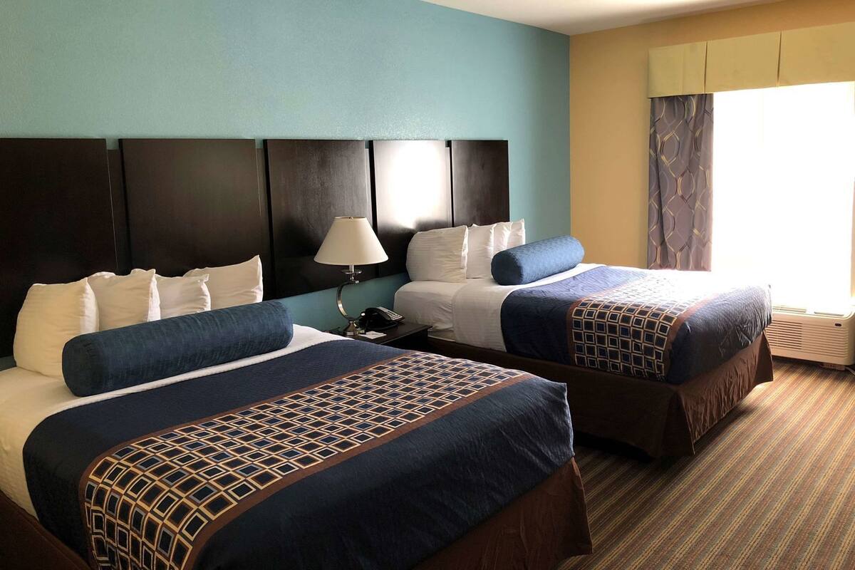 A hotel room with two beds, a headboard, a bedside table with a lamp, and a window with curtains.