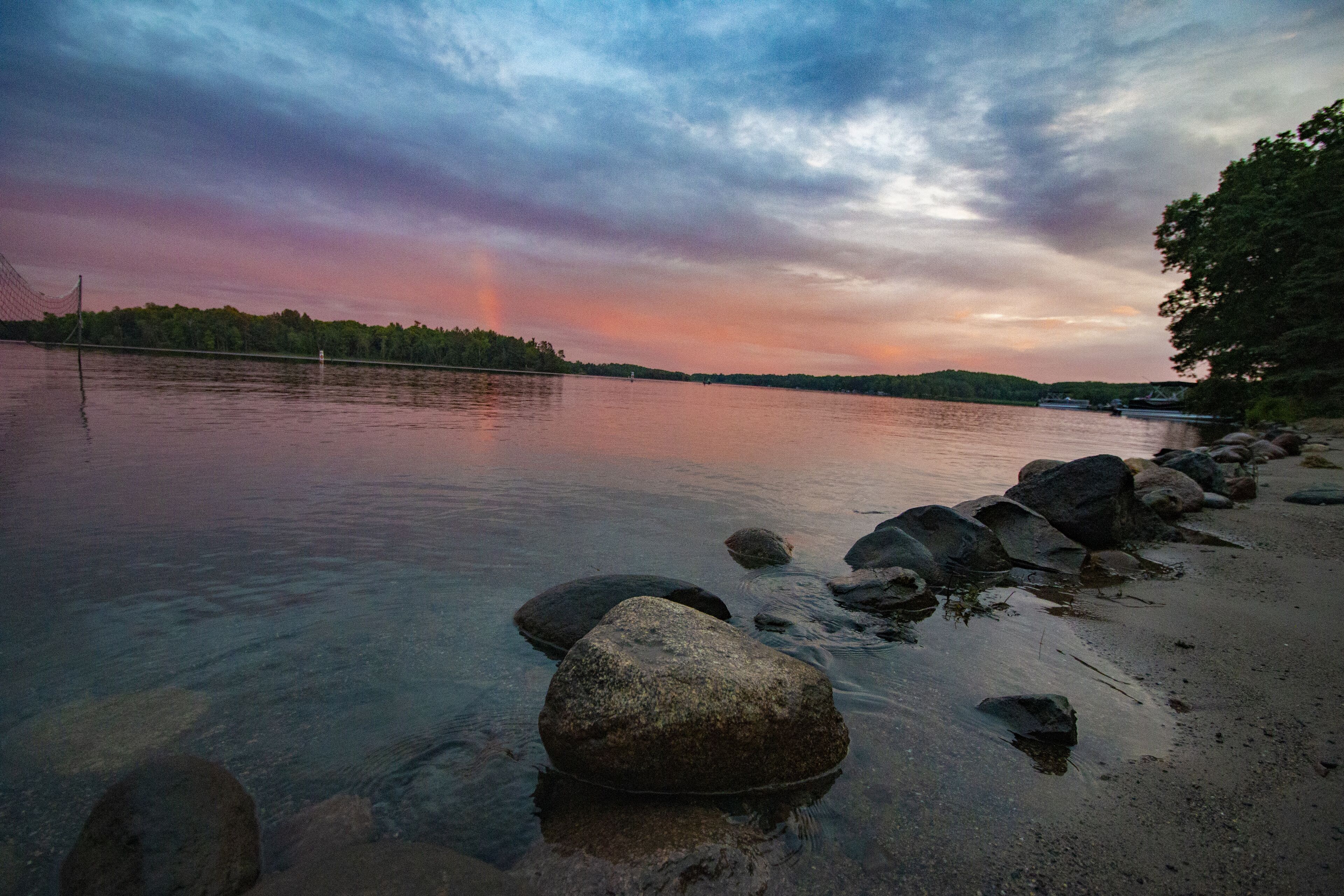 A calm lake with rocks in the foreground and a forested shoreline in the background.