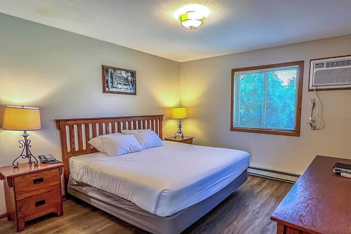 Un dormitorio ordenado con cama, mesitas de noche, lámpara, cuadro enmarcado, ventana con persianas y equipo de aire acondicionado.