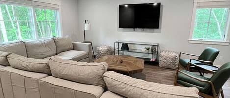 Una sala moderna con un sofá seccional beige grande, un televisor de pantalla plana montado en la pared y dos ventanas con vistas a árboles.