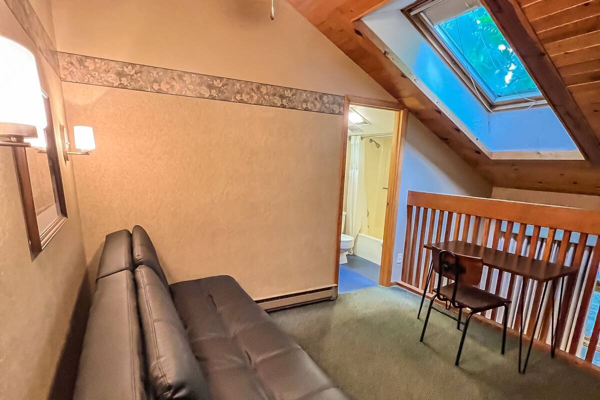 Una habitación con un sofá negro, un ventilador de techo, un tragaluz y una mesa de madera con sillas.