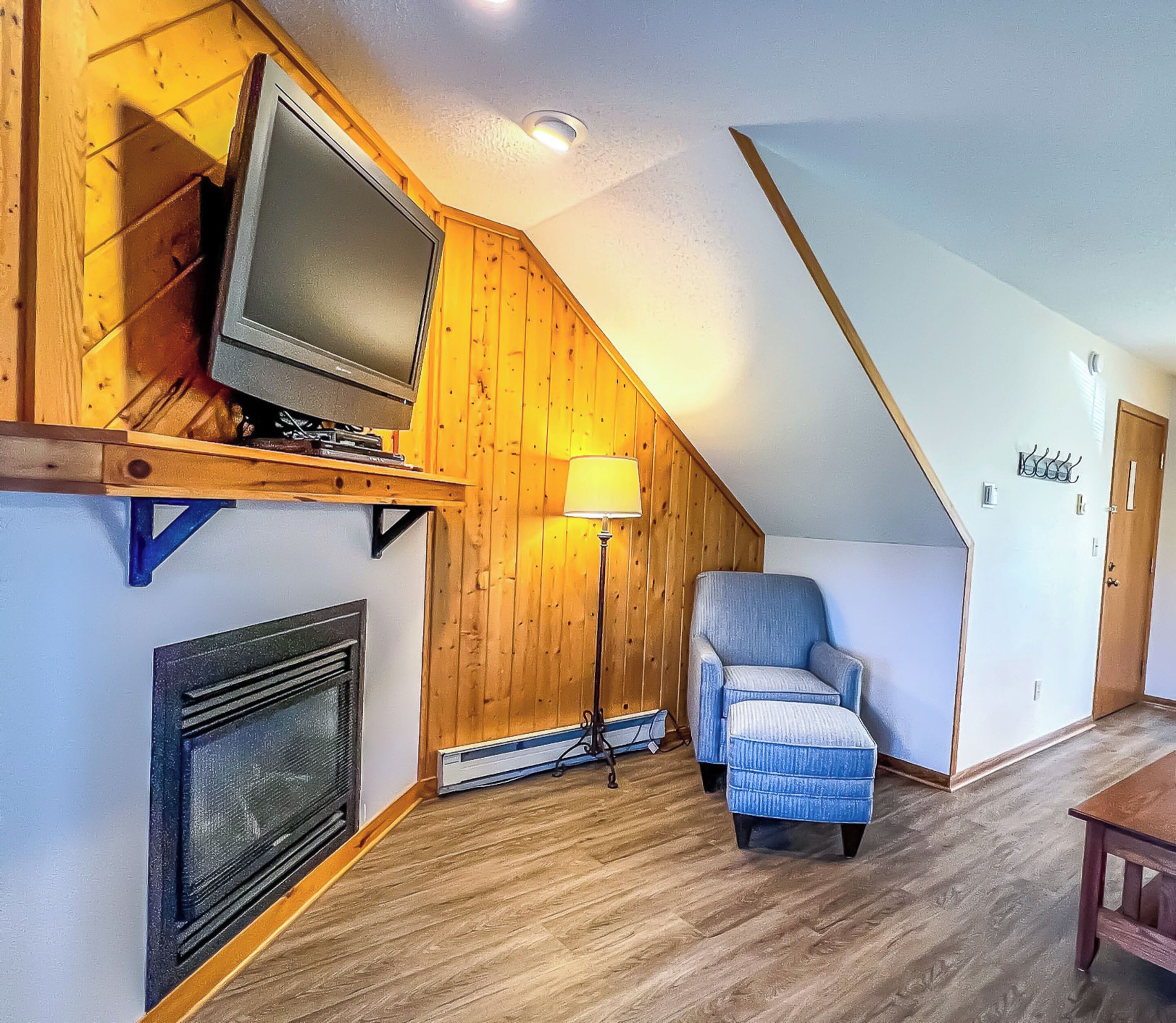 A cozy room with a sloped ceiling, a television on a wooden shelf, a chair, a floor lamp, and a fireplace.