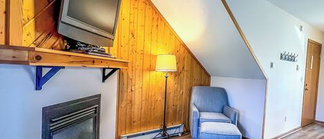 A cozy room with a sloped ceiling, a television on a wooden shelf, a chair, a floor lamp, and a fireplace.