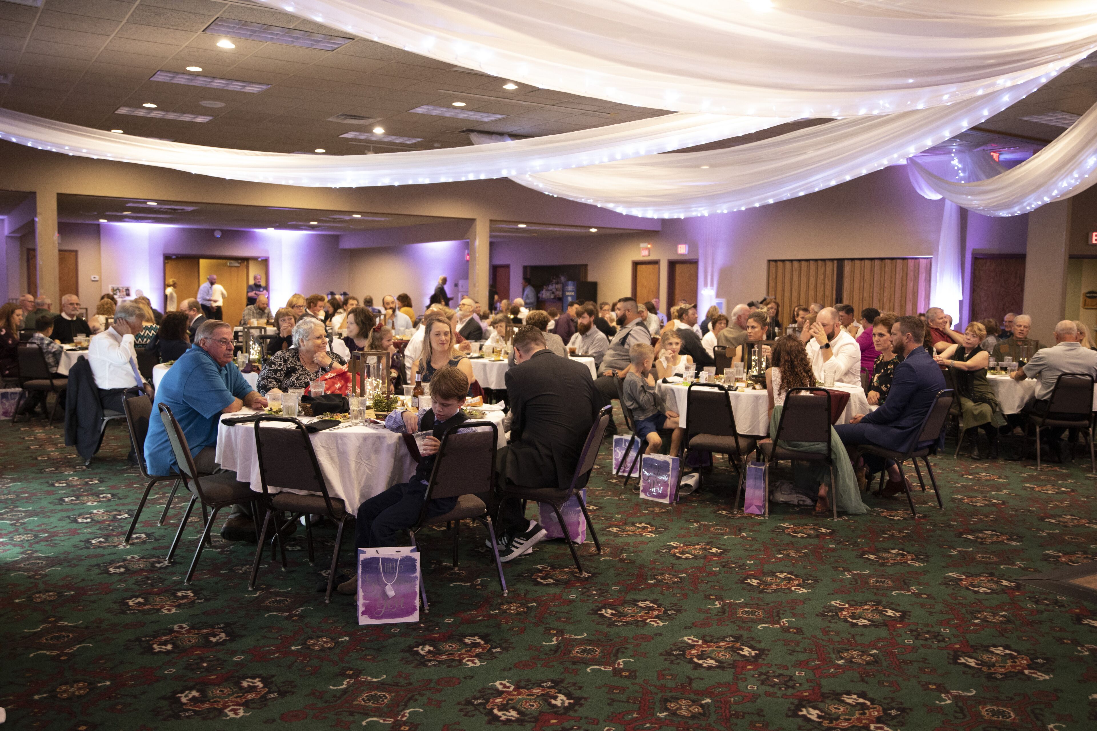 A large indoor event with round tables and chairs, decorated with draped fabric and ambient lighting.
