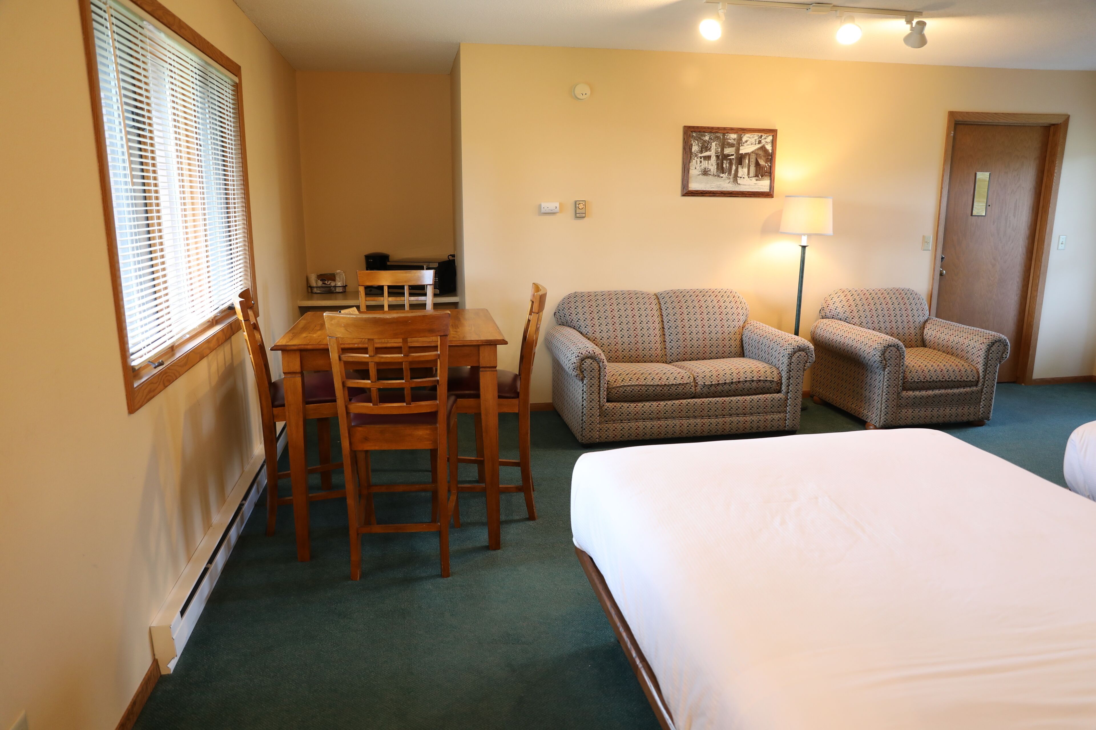 A hotel room with a bed, two armchairs, a wooden table with chairs, a desk, and a lamp.