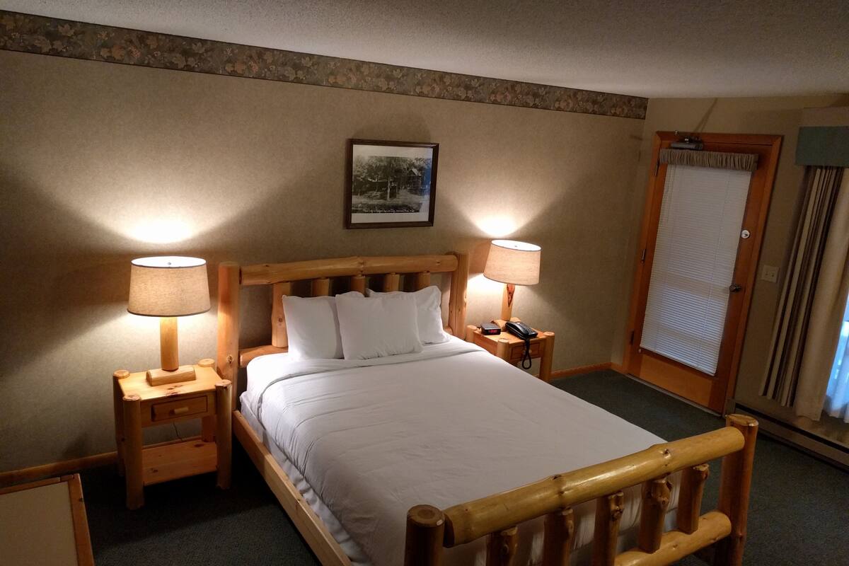 Una cama bien hecha con sábanas blancas, dos lámparas de noche, una mesita de madera y un cuadro en la pared.