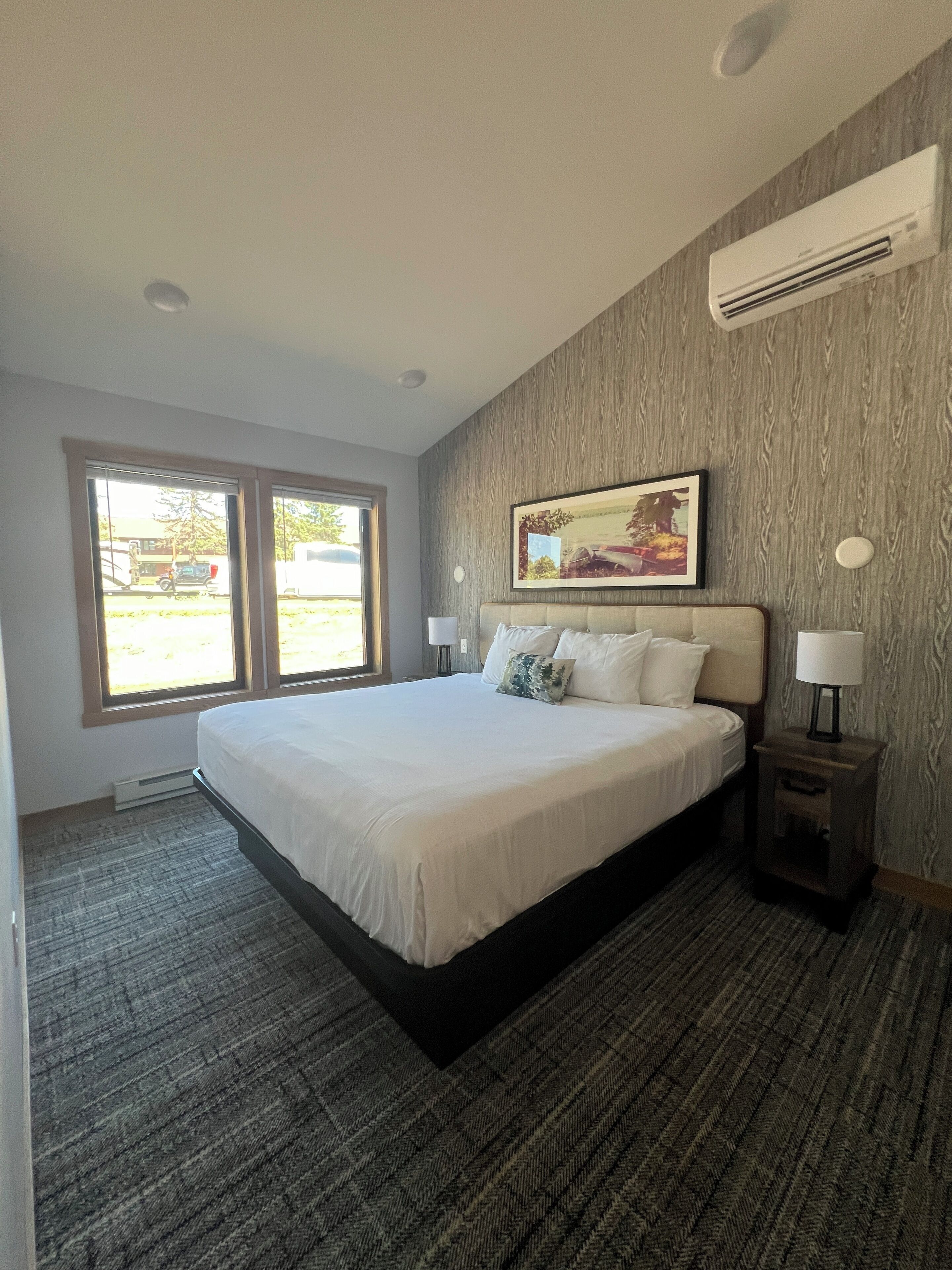 A hotel room with a large bed, a nightstand, a wall-mounted air conditioner, and a window with a view of the outside.