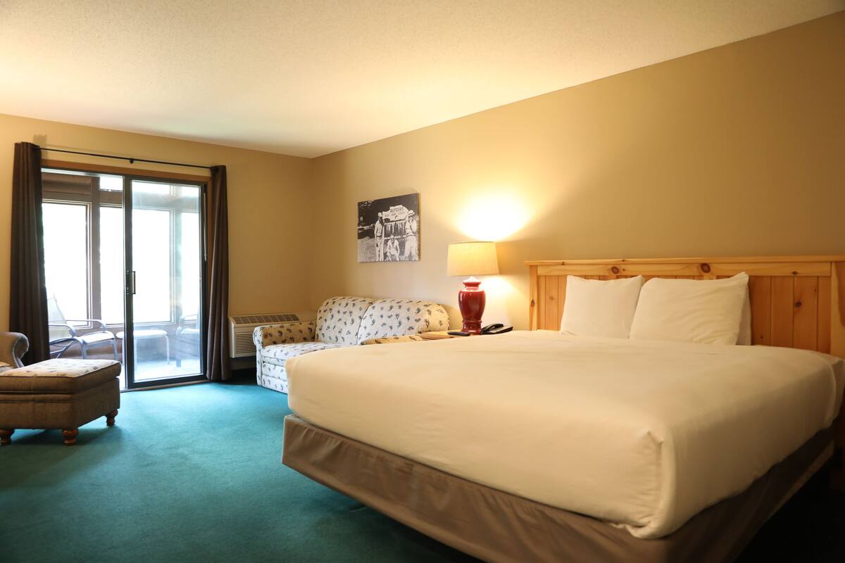 Habitación de hotel con una cama grande, una silla, una lámpara y vistas al exterior a través de una puerta corrediza.