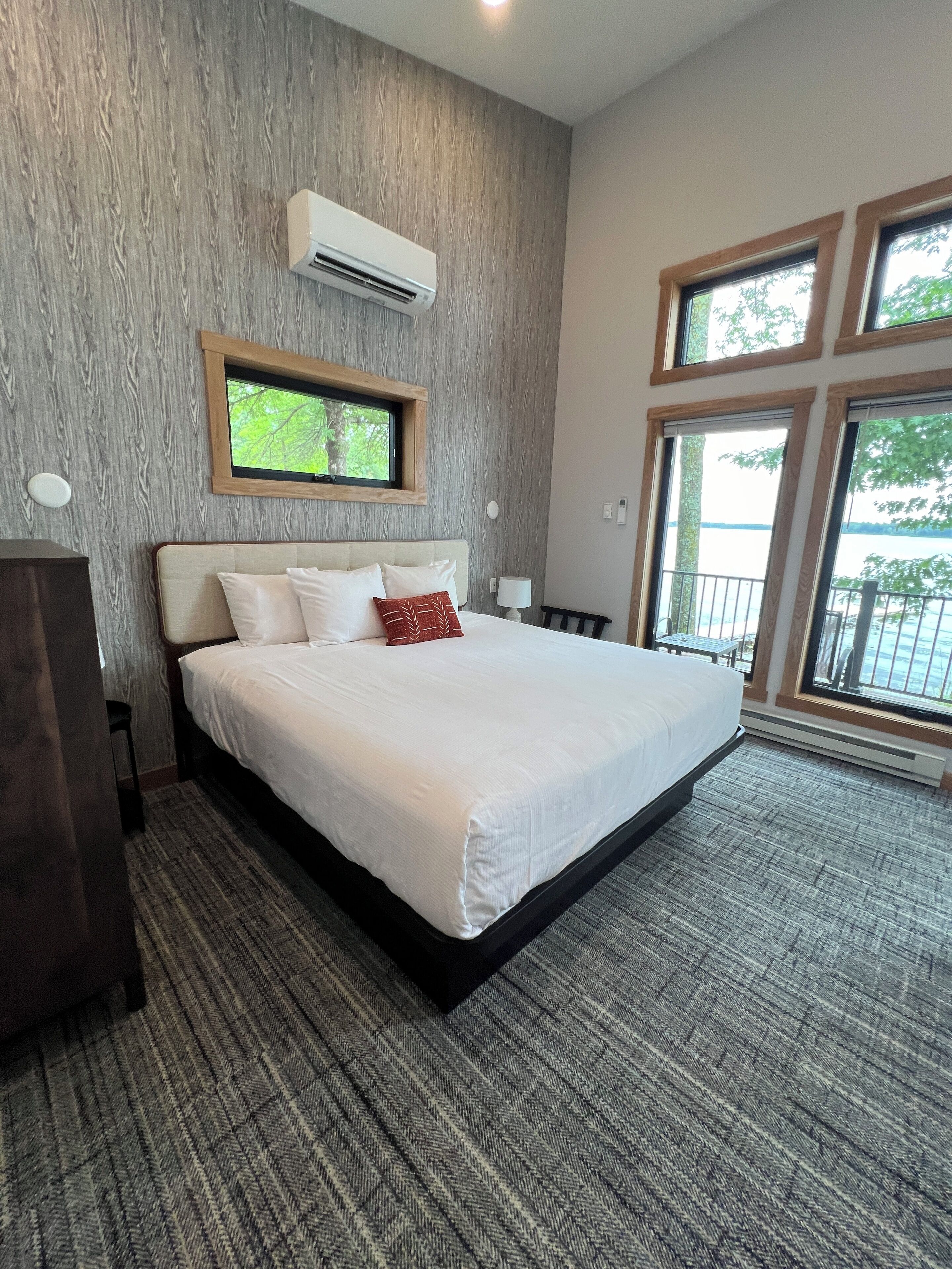 Une chambre d’hôtel moderne dotée d’un grand lit, d’un climatiseur intégré, d’une fenêtre donnant sur un espace verdoyant et d’un balcon avec une balustrade.