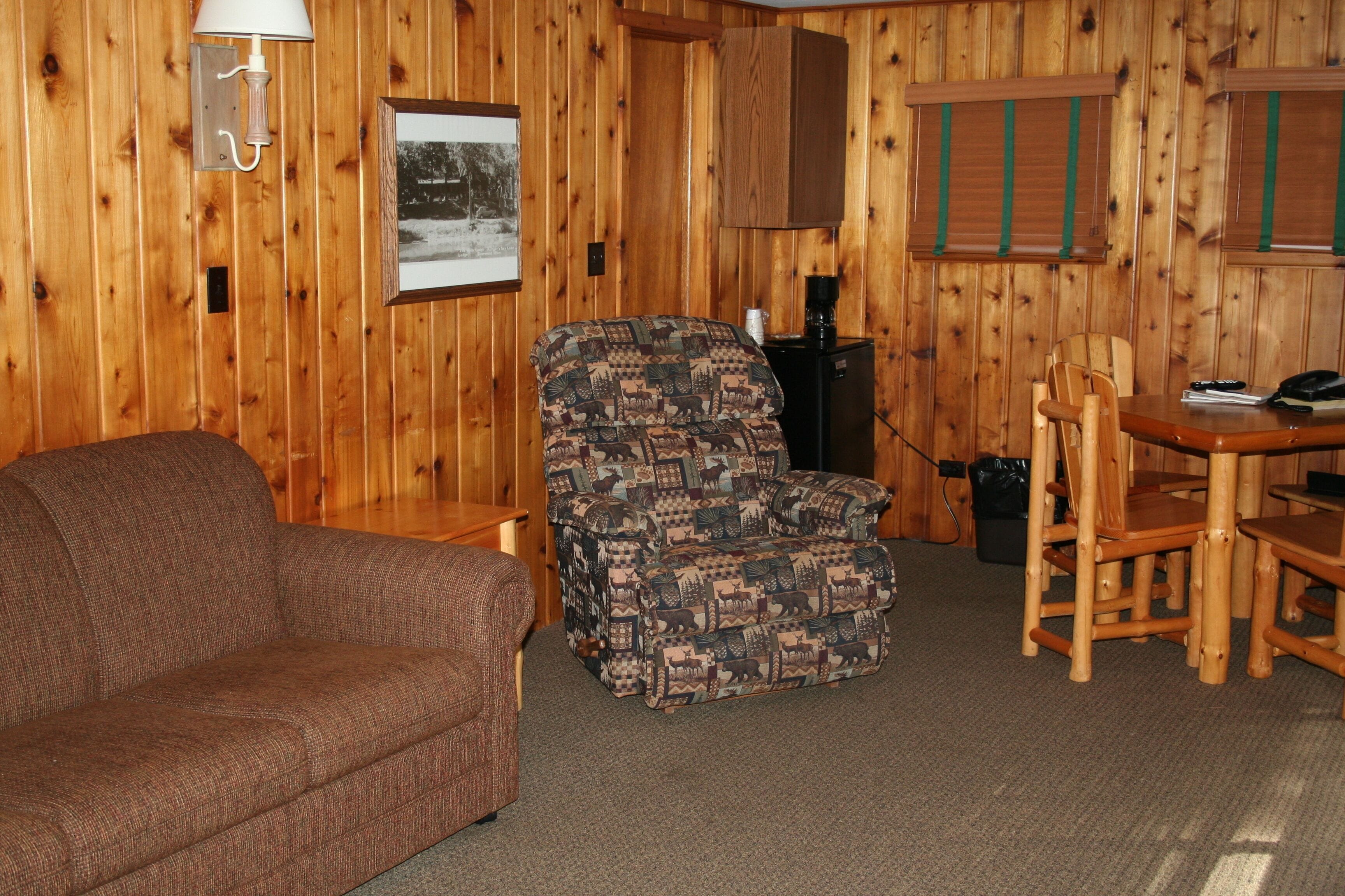 Una habitación acogedora con un sofá marrón, un sillón estampado, una mesa de comedor con sillas, un refrigerador pequeño y una lámpara.