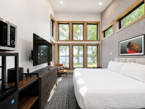 A modern bedroom with a large bed, a TV, a dresser, and a window with a view of trees and water. - Ruttger's Bay Lake Resort (Deerwood)