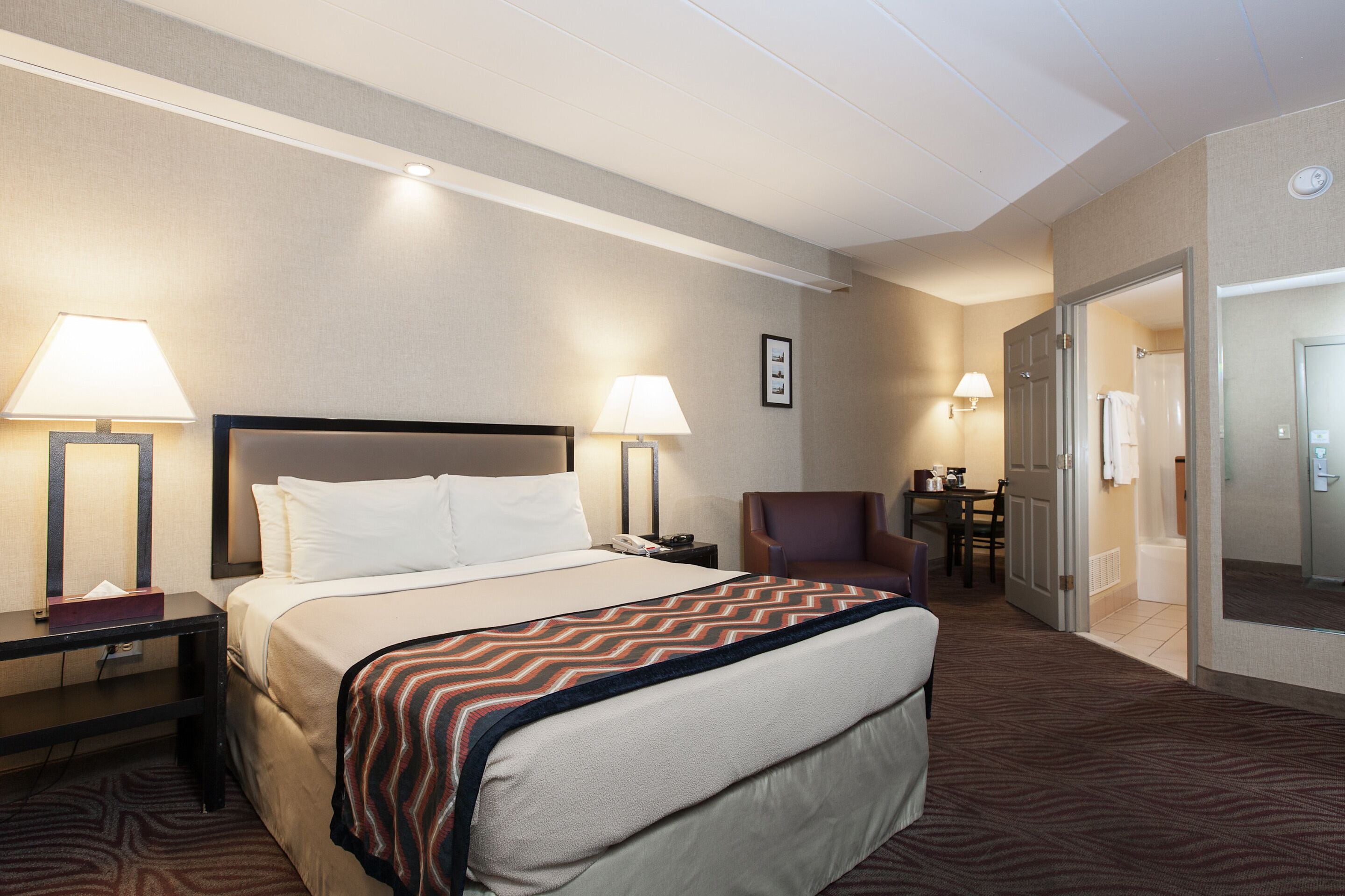 A hotel room with a large bed, two bedside lamps, a chair, a desk, and a door leading to a bathroom.