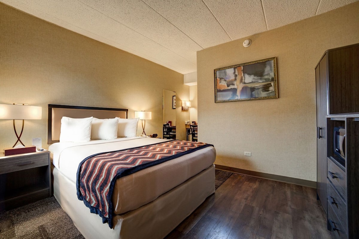A hotel room with a bed, two bedside lamps, a desk with a chair, a television, and a painting on the wall.